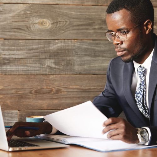 Prosperous top-manager in formal suit working with papers using laptop with internet connection in coffee shop. Young African businessman typing text on computer checking information for job purposes.